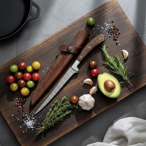 May include: A wooden cutting board with a knife in a leather sheath, surrounded by fresh ingredients. The knife has a wooden handle and a silver blade. Ingredients include tomatoes, garlic, avocado, and herbs. A cast iron pan is in the background.
