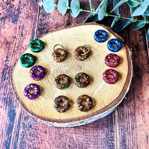 Può includere: Una collezione di orecchini e ciondoli a forma di ciambella, colorati e glitterati. Gli orecchini sono in vari colori, tra cui verde, viola, rosso e blu, e sono esposti su una fetta di legno. Alcuni orecchini hanno cerchietti dorati.