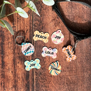 Può includere: Una collezione di targhette identificative colorate per animali domestici in varie forme, tra cui disegni a forma di osso e circolari. Ogni targhetta presenta un nome unico come "Candy", "Peach", "Joy", "Lola", "Pinky" e "Sunny" in un carattere giocoso, con motivi a scacchi e floreali.