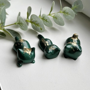 Peut inclure: Une collection de petites figurines en céramique vert foncé avec des accents de feuilles d'or. Les figurines comprennent un chat, deux objets en forme de bouteille et deux formes arrondies. Les objets sont disposés sur une surface blanche, avec des feuilles d'eucalyptus en arrière-plan.