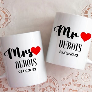 Peut inclure: Deux tasses en céramique blanche avec du texte noir et des cœurs rouges. La tasse de gauche dit "Mme DUBOIS 23.09.2022" et la tasse de droite dit "M. DUBOIS 23.09.2022".