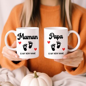 Peut inclure: Deux mugs en céramique blancs avec du texte et des graphiques noirs. L'un des mugs dit "Maman" et l'autre "Papa". Les deux mugs ont des empreintes de pieds de bébé noires et des cœurs rouges. Le texte "IS MY NEW NAME" est en dessous.
