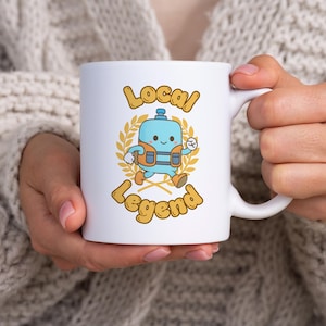 Peut inclure: Mug en céramique blanche avec l'inscription "Local Legend" et un personnage de dessin animé. Le personnage est bleu clair avec un gilet orange et court. Le texte et le personnage sont entourés d'une couronne dorée.