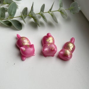 Peut inclure: Trois figurines roses en forme de chat avec des accents de feuilles d'or. Les figurines sont dans différentes poses, dont une couchée et les autres debout. L'arrière-plan comprend une branche de feuilles vertes sur une surface blanche.