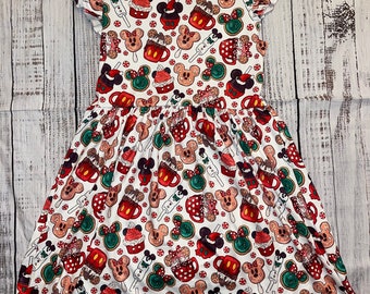 Pode incluir: Vestido branco com um padrão festivo com biscoitos de gengibre, chocolate quente e picolés com o tema Mickey Mouse em vermelho, verde e marrom. O vestido tem mangas curtas com babados e uma saia franzida.