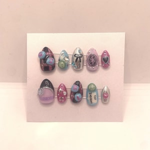 May include: A set of ten artificial nails in various designs. The nails feature a mix of colours, including pink, black, silver, and blue, with embellishments like bows, stars, and small round accents. Some nails have text, and others have heart and star shapes.