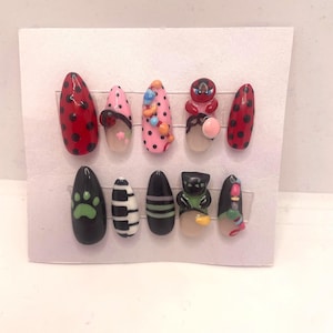 May include: A set of ten artificial nails with various designs. The nails feature a mix of colors, including red with black dots, pink with black dots, and black with green paw prints. Some nails have 3D embellishments.