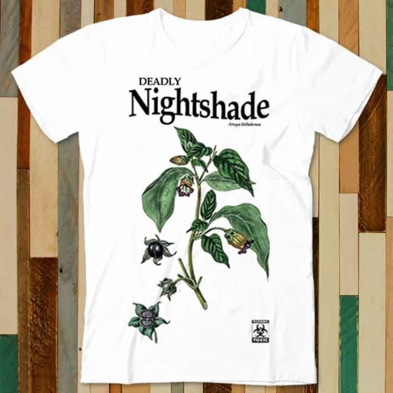 May include: White t-shirt featuring a black and green illustration of a deadly nightshade plant. The text "DEADLY NIGHTSHADE" is printed in black above the plant. The text "Atropa Belladonna" is printed in smaller black text below the plant. A black and white warning sign with the text "DANGER TOXIC" is printed in the bottom right corner of the shirt.