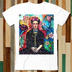 Puede incluir: Una camiseta blanca con un estampado gráfico colorido de Frida Kahlo rodeada de flores y un pájaro azul. El gráfico es una ilustración vibrante y detallada de la famosa artista mexicana.