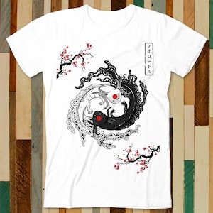 Puede incluir: Camiseta blanca con un diseño de ajolote en blanco y negro al estilo yin-yang. El diseño incluye detalles en rojo y texto japonés. Ramas de flores de cerezo enmarcan los ajolotes.