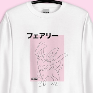 Puede incluir: Sudadera blanca con un dibujo lineal negro de un personaje Sylveon sobre un rectángulo rosa. La palabra japonesa "Fairy" está encima del gráfico. La sudadera está colgada de una percha negra.