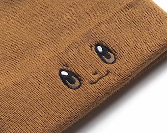 May include: A brown knitted beanie with a cute embroidered face. The face has two large black eyes and a small smile.