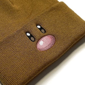 Può includere: Un berretto di lana marrone con un viso di cartone animato ricamato sul davanti. Il viso ha due occhi neri, un naso nero e una bocca rosa.