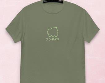 May include: Olive green t-shirt with a neon green outline of a creature and Japanese text. The t-shirt is hanging on a black hanger against a pink background. The short-sleeved shirt is made of a soft material.