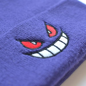 May include: A purple knitted beanie with an embroidered Gengar face. The design features red eyes, a white grin, and black outlines. The beanie is made of a soft, textured material, ideal for colder weather.