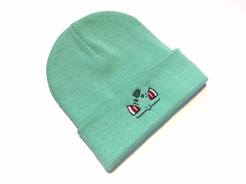 May include: A light teal beanie hat with a turned-up hem. The hat has an embroidered Bulbasaur face in black, red, and green. The beanie is made of a soft, knitted material. A fun accessory for fans of the popular franchise, ideal for keeping warm in winter.