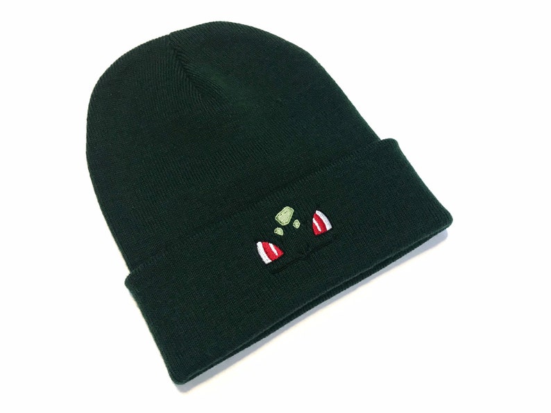 May include: Dark green beanie hat with a turned-up hem. The hat has an embroidered cartoon character face in green, red, and white. The beanie is made of a soft, knitted fabric.
