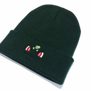 May include: A dark green knitted beanie hat with a turned-up brim. The front of the hat has an embroidered cartoon face design, with red and white details. The hat is made of a soft, knitted material.