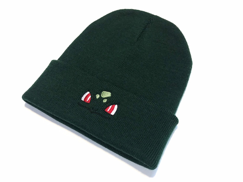 May include: A dark green knitted beanie hat with a turned-up brim. The front of the hat has an embroidered cartoon face design, with red and white details. The hat is made of a soft, knitted material.