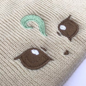 May include: Beige knit beanie with an embroidered cartoon face. The face features brown eyes with white highlights and a light green swirl above. The beanie is made of a soft, textured material.