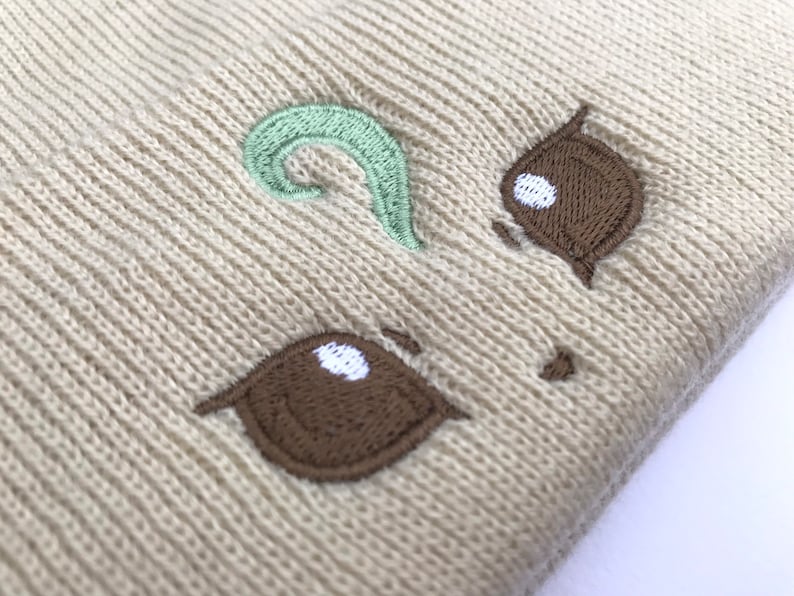May include: Beige knit beanie with an embroidered cartoon face. The face features brown eyes with white highlights and a light green swirl above. The beanie is made of a soft, textured material.