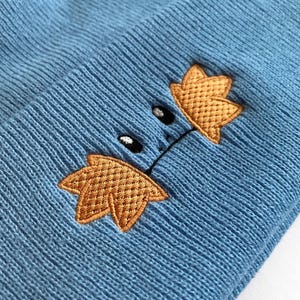 Op de afbeelding: Een lichtblauwe gebreide muts met een geborduurd cartoongezicht. Het ontwerp heeft twee goudgele bladvormen en zwarte ogen. De muts is gemaakt van een zacht, getextureerd materiaal.