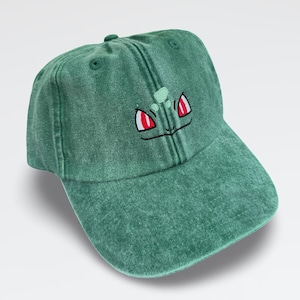 May include: A green baseball cap with a washed, distressed texture. The front panel features an embroidered graphic of two red eyes with white highlights and a small light green shape above them, resembling a stylised creature face.