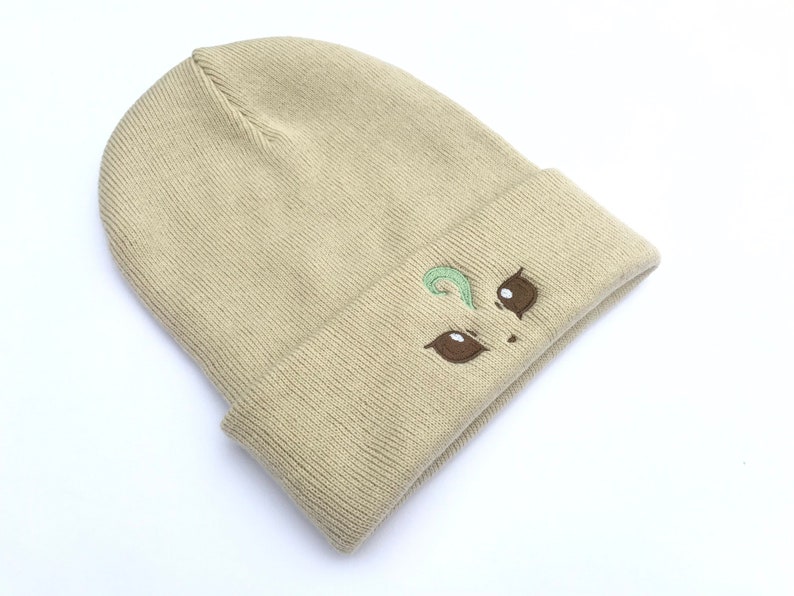 May include: Tan beanie hat with a folded brim. The hat features an embroidered design of two brown eyes and a green swirl. The hat is made of a soft, knitted material and is suitable for cold weather.