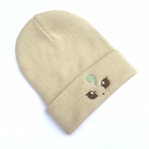 May include: Tan knit beanie with an embroidered cartoon face design. The design features brown eyes and a green swirl above the eyes. The beanie is folded at the brim and is made of a soft, knitted material.