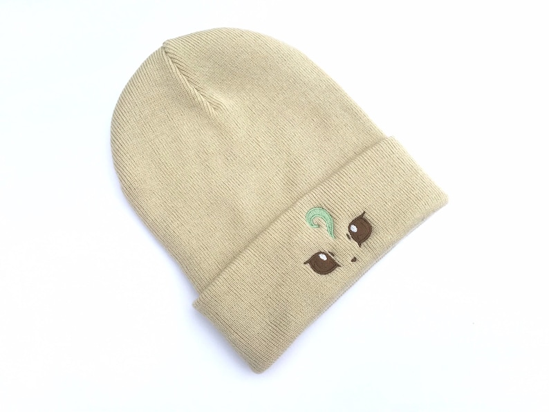 May include: Tan knit beanie with an embroidered cartoon face design. The design features brown eyes and a green swirl above the eyes. The beanie is folded at the brim and is made of a soft, knitted material.