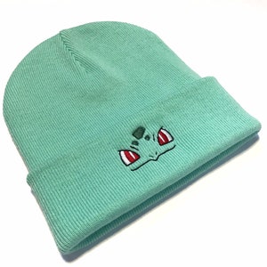 May include: A light teal beanie hat with a turned-up brim. The front of the hat has an embroidered cartoon character face design, with red and white eyes and a green face.