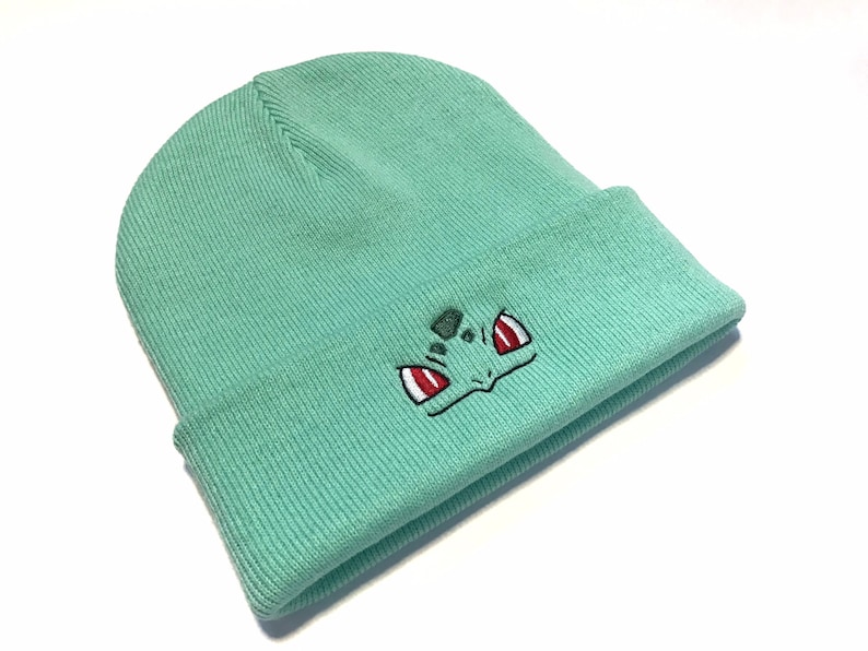 May include: A light teal beanie hat with a turned-up brim. The front of the hat has an embroidered cartoon character face design, with red and white eyes and a green face.