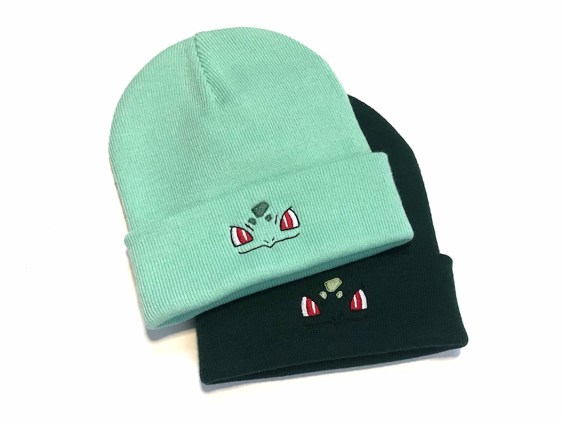 May include: Two knitted beanies, one in light teal and the other in dark green. Each beanie has an embroidered cartoon character face on the folded brim. The design includes red, white, and green details.