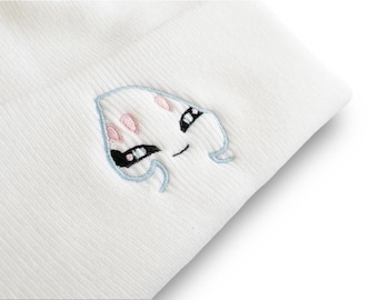 May include: White beanie with an embroidered cartoon ghost design. The ghost has black eyes, pink accents, and a light blue outline. The beanie is made of a ribbed knit fabric.