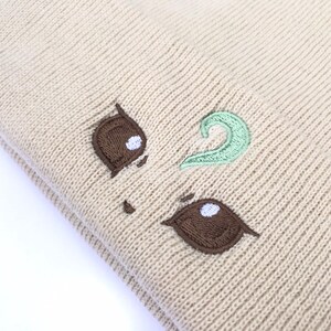 May include: Beige knit beanie with embroidered brown and mint green details. The beanie features a whimsical design with brown eye shapes and a mint green swirl, creating a playful aesthetic. The hat is made of a soft, knitted material.