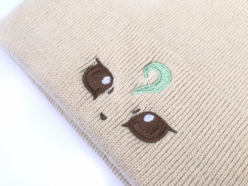 May include: Beige knit beanie with embroidered brown and mint green details. The beanie features a whimsical design with brown eye shapes and a mint green swirl, creating a playful aesthetic. The hat is made of a soft, knitted material.