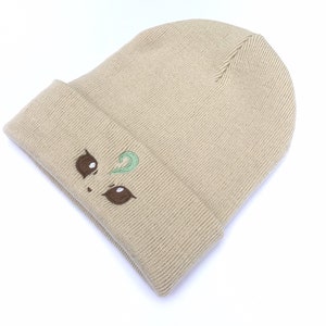May include: Tan knit beanie with a folded brim. The beanie features an embroidered face design with brown eyes and a green swirl. The hat is made of a soft, ribbed knit material.