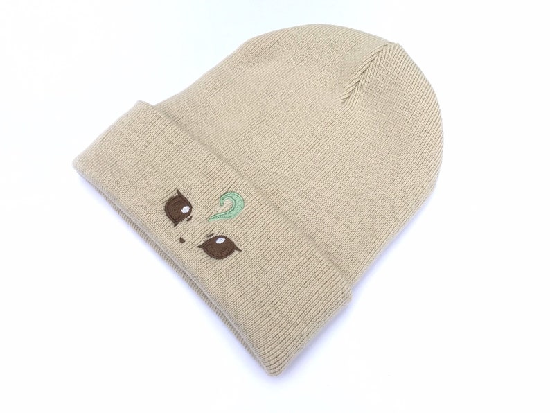 May include: Tan knit beanie with a folded brim. The beanie features an embroidered face design with brown eyes and a green swirl. The hat is made of a soft, ribbed knit material.