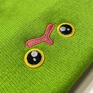 May include: Lime green knitted beanie hat with embroidered cartoon eyes and nose. The eyes have black pupils with yellow edging. The nose is pink. A playful accessory.