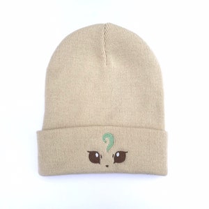 May include: A tan beanie hat with a folded brim. The hat features an embroidered design of a cartoon face with brown eyes and a green question mark above the eyes. The hat is made of a knitted material.