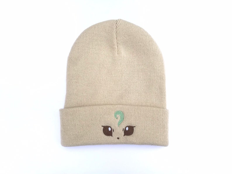 May include: A tan beanie hat with a folded brim. The hat features an embroidered design of a cartoon face with brown eyes and a green question mark above the eyes. The hat is made of a knitted material.
