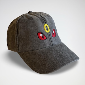 Può includere: Un cappellino da baseball grigio con visiera curva. Il cappellino presenta ricami di occhi rossi e gialli. Il cappello è realizzato in tessuto strutturato e ha un bottone sulla parte superiore.