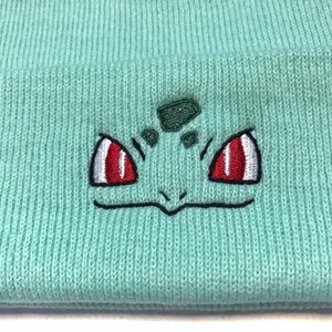 May include: A light teal knitted beanie hat with an embroidered Bulbasaur design. The Bulbasaur face has red and white eyes, a green head, and a black outline. The hat is made of a soft, ribbed knit material.