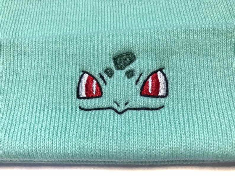 May include: A light teal knitted beanie hat with an embroidered Bulbasaur design. The Bulbasaur face has red and white eyes, a green head, and a black outline. The hat is made of a soft, ribbed knit material.