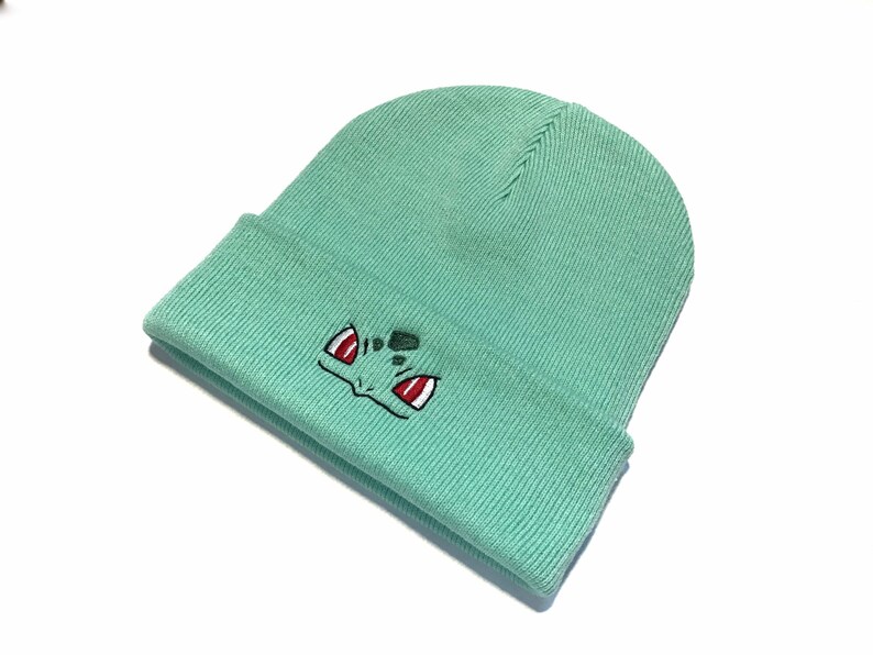 May include: A pale green beanie hat with a turned-up hem. The hat has an embroidered cartoon face in black, red, and green. The beanie is made of a ribbed knit fabric.