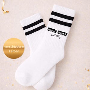 Könnte beinhalten: Ein Paar weiße Socken mit schwarzen Streifen und dem Text "COOLE SOCKE seit 1986". Im Hintergrund befindet sich ein goldener Kreis mit dem Text "verschiedene Farben".