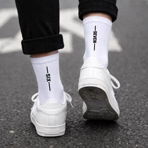 Könnte beinhalten: Ein Paar weiße Socken mit schwarzem Text, der vertikal auf der Außenseite jeder Socke aufgedruckt ist. Die linke Socke zeigt das Wort SIX zwischen zwei horizontalen Linien, die rechte Socke zeigt das Wort SEVEN zwischen zwei Linien.