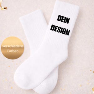 Könnte beinhalten: Ein Paar weiße Socken mit den Wörtern "DEIN DESIGN" in Schwarz gedruckt. Ein goldener Kreis mit den Worten "verschiedene Farben" befindet sich in der unteren linken Ecke. Die Socken befinden sich auf einem hellrosa Hintergrund.