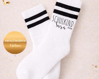 Puede incluir: Un par de calcetines blancos con dos rayas negras horizontales en la parte superior. El calcetín derecho muestra el texto negro SCHULKIND Sara 2026. Un círculo dorado a la izquierda contiene el texto blanco verschiedene Farben.