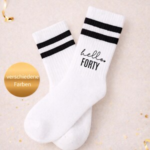 Könnte beinhalten: Ein Paar weiße Socken mit zwei horizontalen schwarzen Streifen am oberen Bündchen. Auf der rechten Socke steht in schwarzer Schrift "hello" in Schreibschrift über dem Wort "FORTY" in einer fetten serifenlosen Schrift mit einem kleinen Herz.
